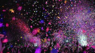 Buntes Konfetti und kleine glitzernde Sterne regnen auf eine Menschenmenge herab, die bei einem nächtlichen Event oder Festival ihre Arme begeistert in die Höhe streckt. Die Szene wird durch farbenfrohe Lichter in Pink, Blau und Gelb beleuchtet.